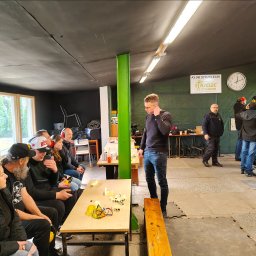 Szkoła Ochrony Kurs Ochrony STS Security Tactical Services Skierniewice - Instruktor prowadzi szkolenie strzeleckie w klubie, uczestnicy siedzą przy stołach z ochronnikami słuchu i okularami. W tle widoczny zegar i logo klubu strzeleckiego.