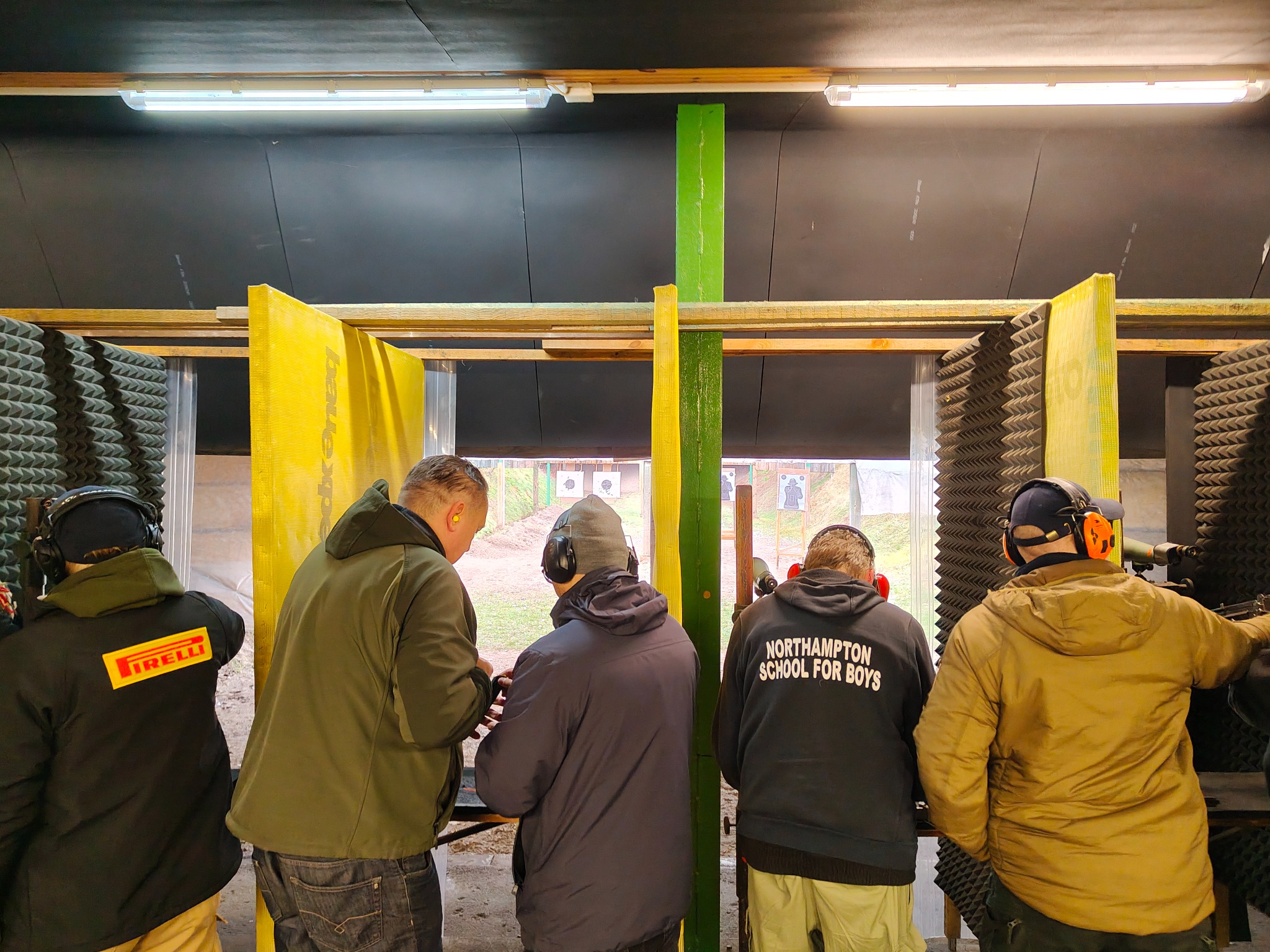 Strzelnica: grupa osób w słuchawkach na strzelnicy, celują do tarcz. Żółte i zielone przegrody, wygłuszenie ścian. Osoby ubrane w kurtki z napisami.
