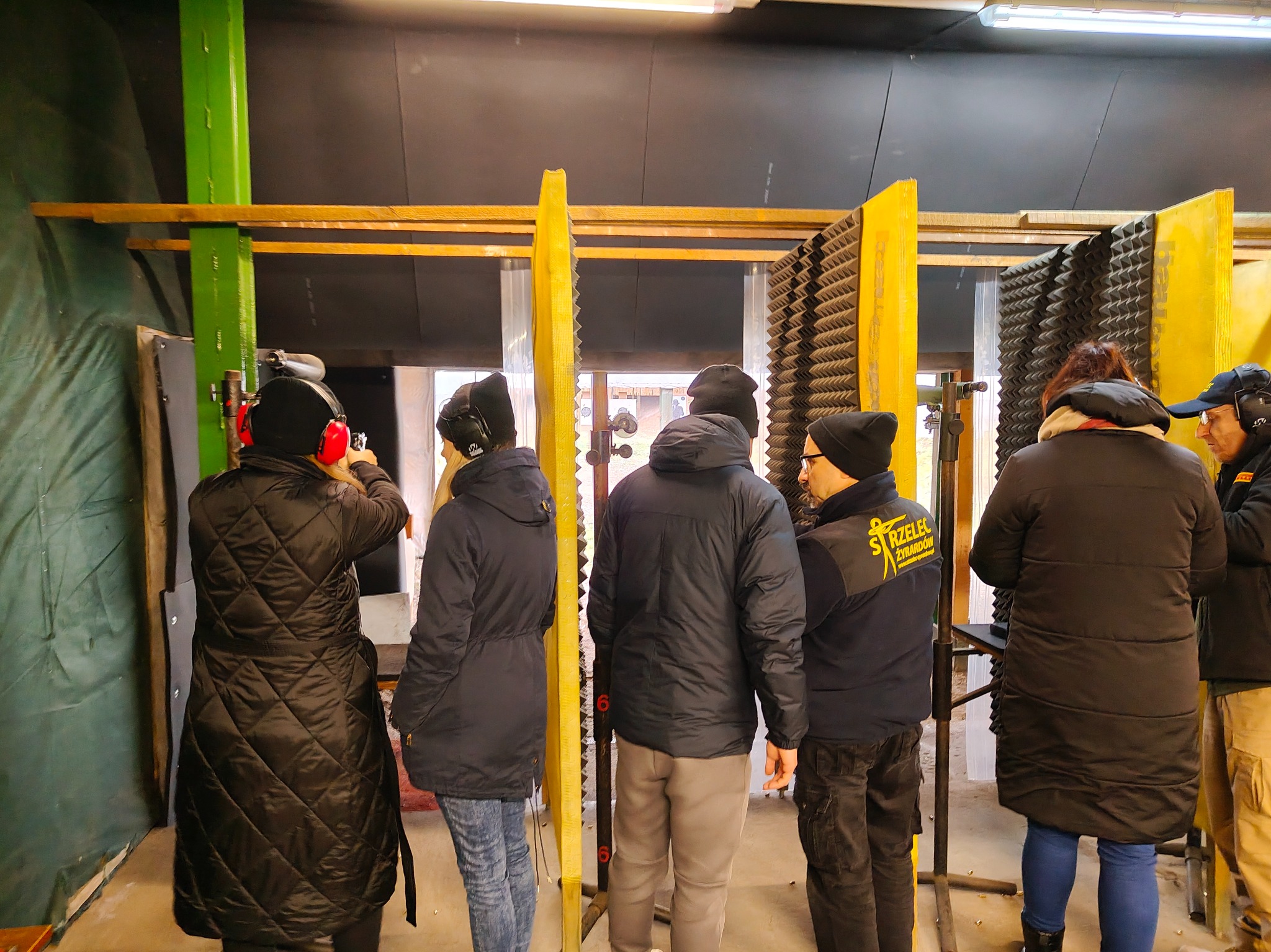 Grupa osób w zimowych kurtkach na strzelnicy, jedna osoba w słuchawkach strzela z pistoletu, żółte przegrody dźwiękochłonne, widok z tyłu.