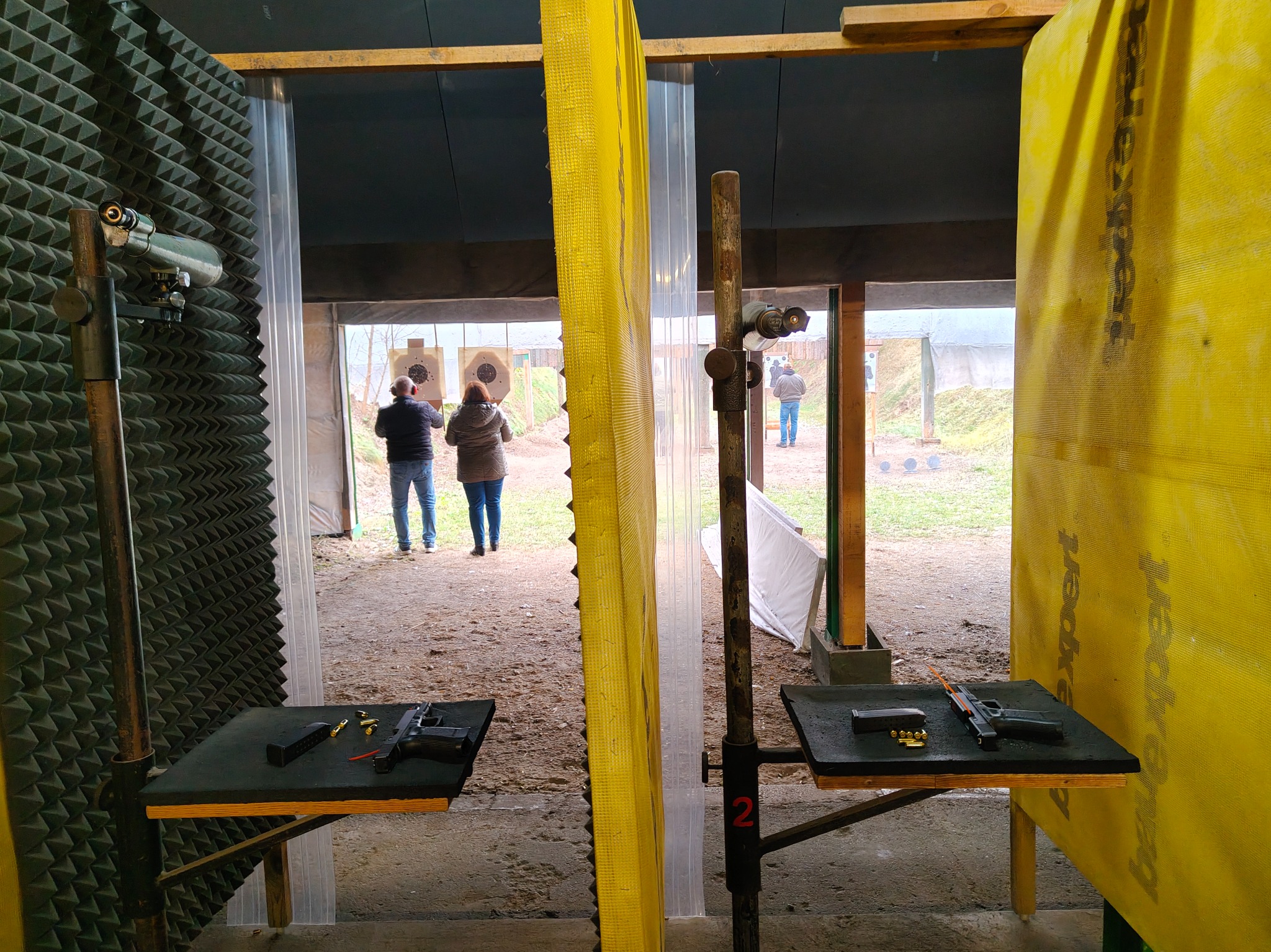 Strzelnica: osoby strzelają do tarcz, na stolikach broń i amunicja. Widok zza stanowiska, z żółtą przegrodą i numerem 2. Akustyczne panele na ścianach.