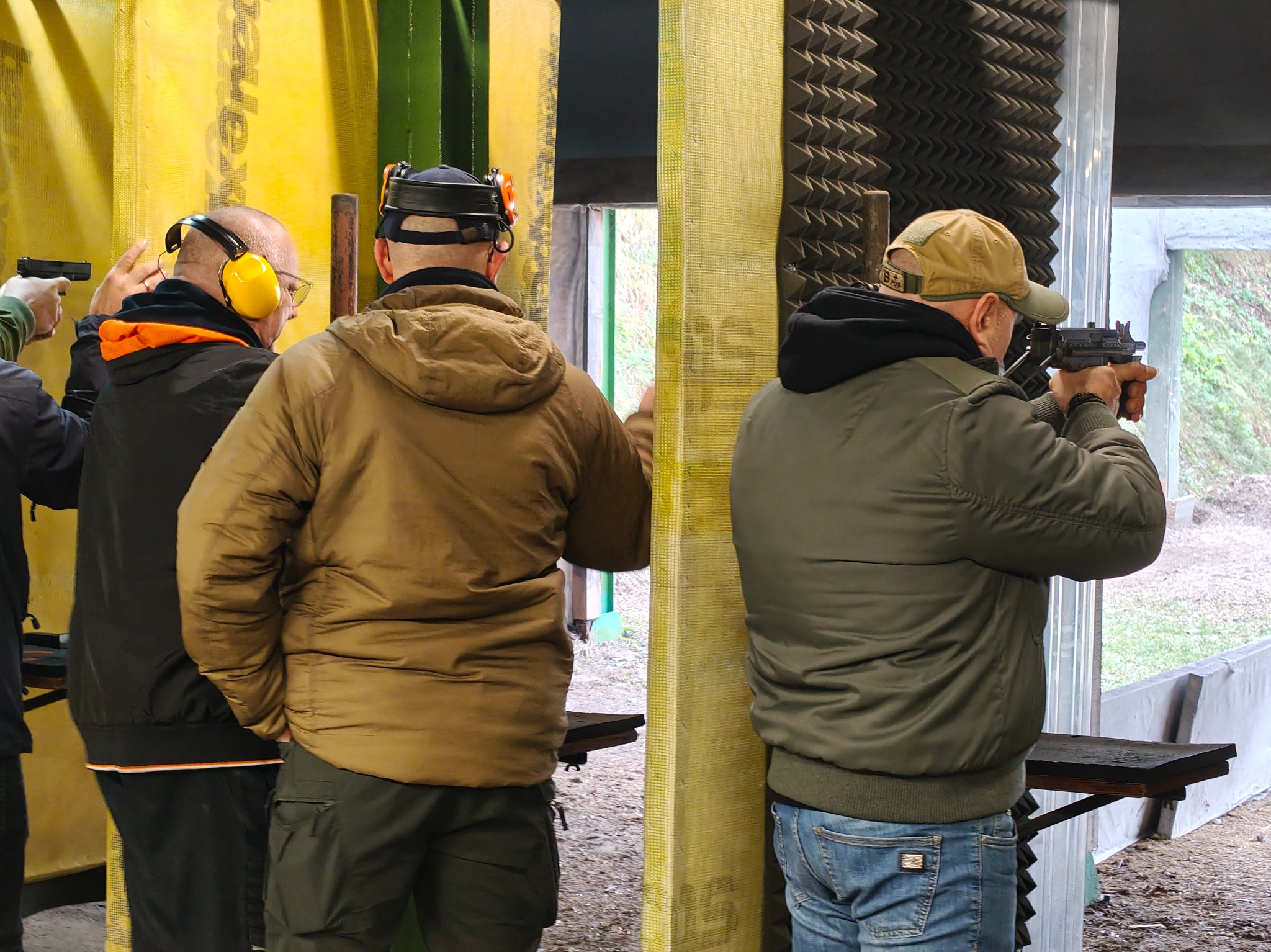 Strzelnica: osoby w słuchawkach i okularach ochronnych ćwiczą strzelanie z broni palnej do tarczy. Widoczne stanowiska strzeleckie i wygłuszone ściany.