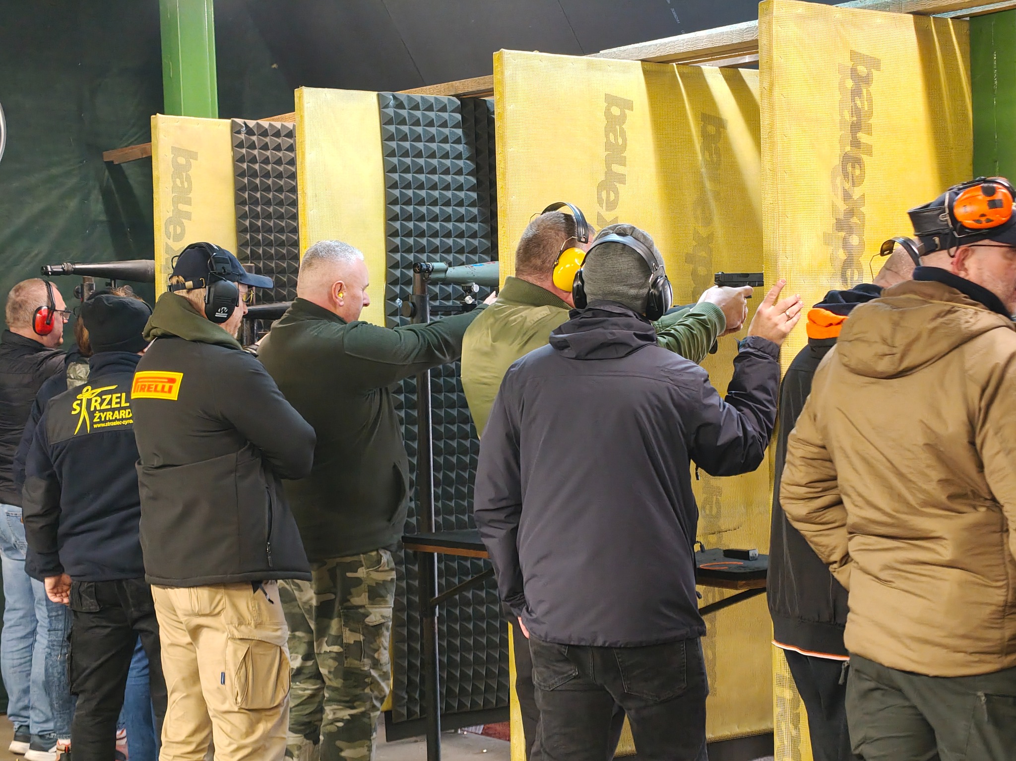 Strzelnica kryta: grupa osób w słuchawkach ochronnych, ćwiczących strzelanie z broni palnej do tarczy. Widoczne stanowiska strzeleckie i broń.