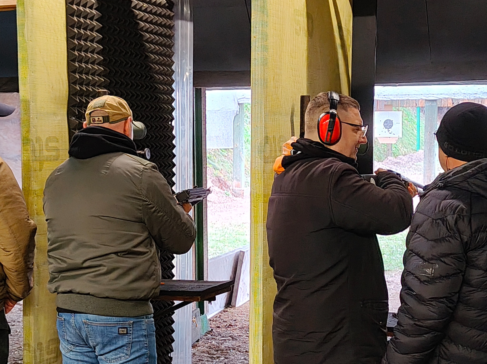 Strzelnica: mężczyźni w ochronnikach słuchu i okularach strzelają do tarczy. Widoczny cel z przestrzelinami, co sugeruje trening strzelecki.