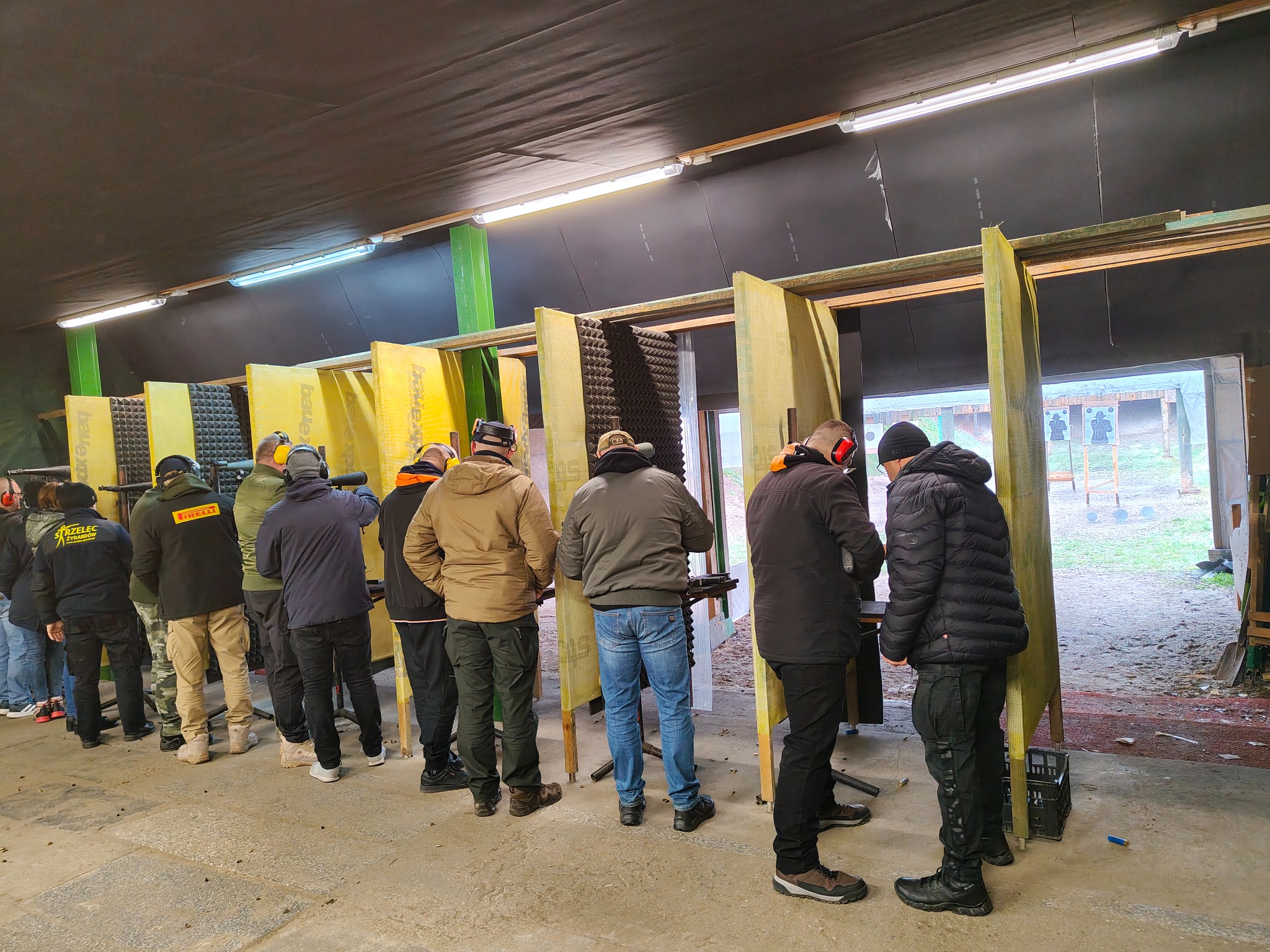 Grupa osób na strzelnicy krytej, w trakcie treningu strzeleckiego. Widoczne tarcze, stanowiska i osoby w słuchawkach ochronnych.