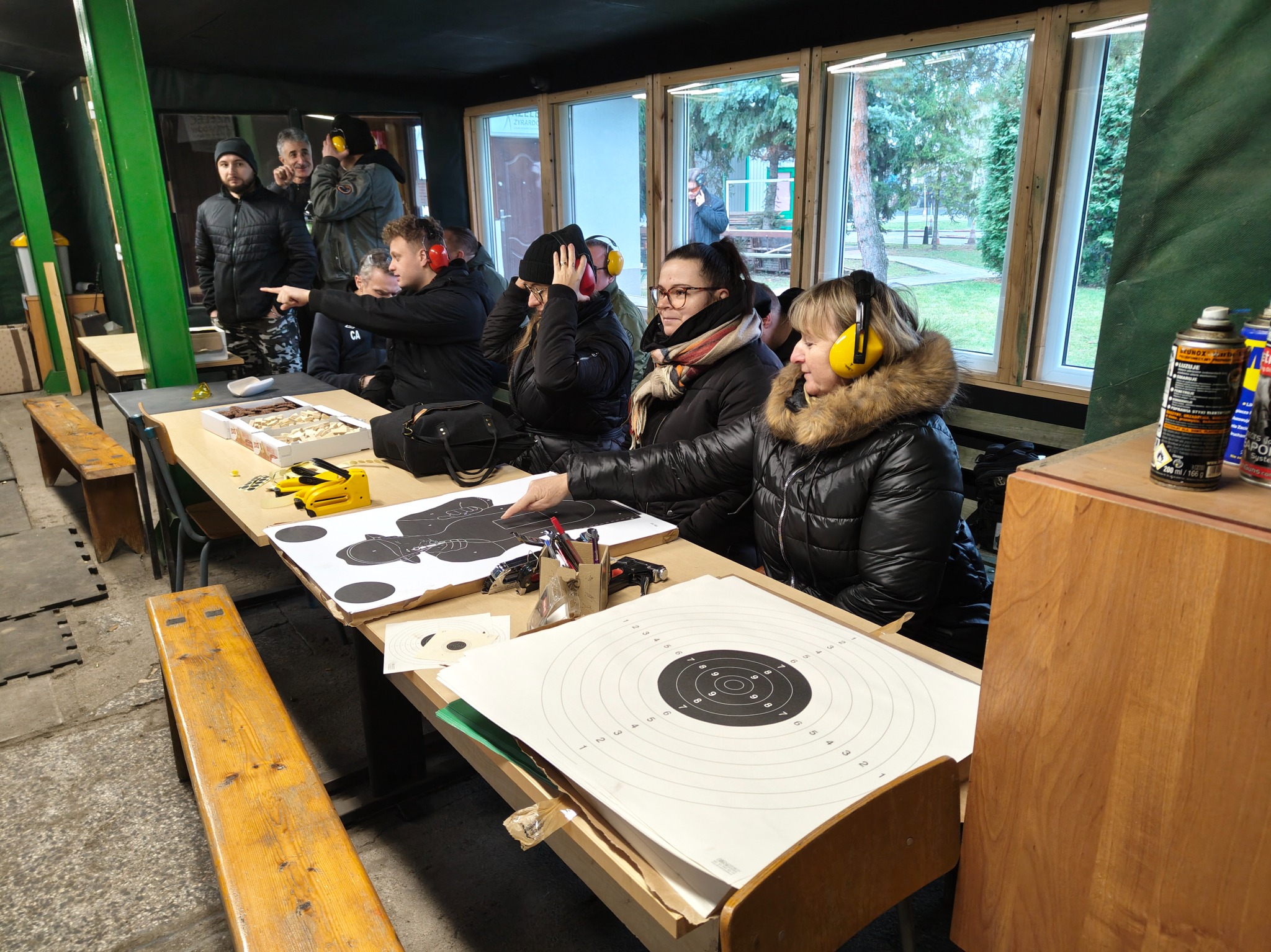 Uczestnicy kursu strzeleckiego w słuchawkach ochronnych, siedzący przy stole z tarczami i przyborami, w tle instruktorzy i okna z widokiem na zewnątrz.