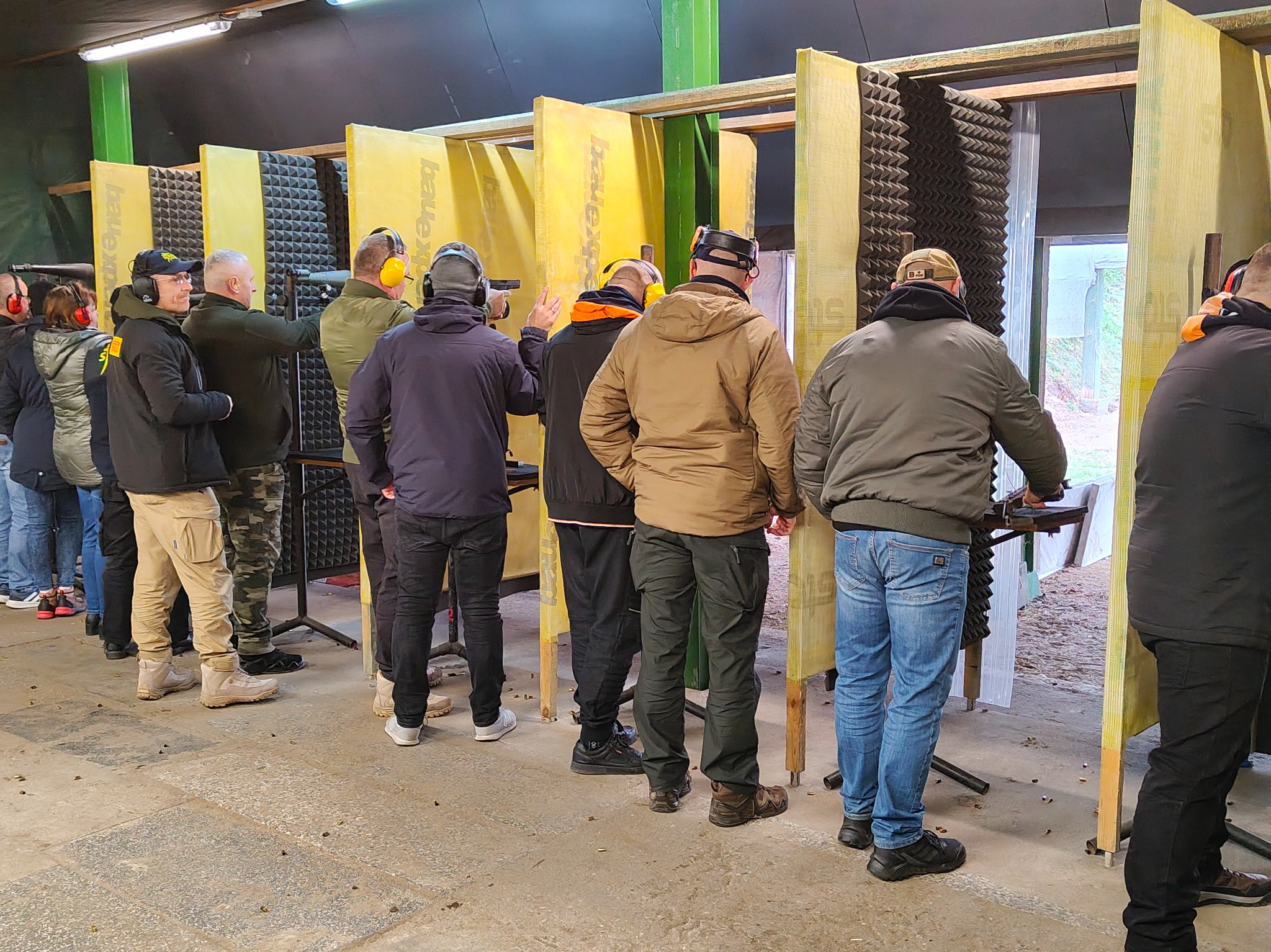 Grupa osób w słuchawkach na strzelnicy, celuje z broni do tarczy. Żółte przegrody oddzielają stanowiska. Widoczne ślady po kulach na podłodze.
