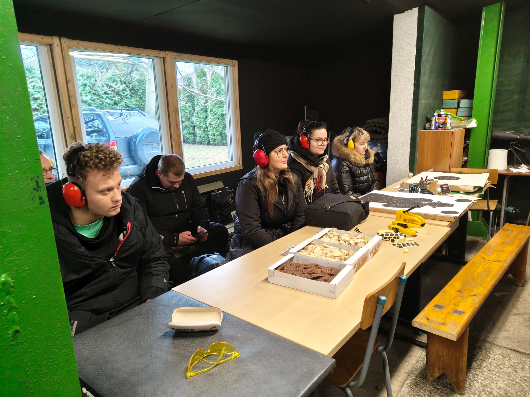 Grupa osób w słuchawkach ochronnych siedzi przy stole w strzelnicy, obok tarcze i przekąski. Widoczne okno z widokiem na zewnątrz, sugerujące lokalizację w budynku.