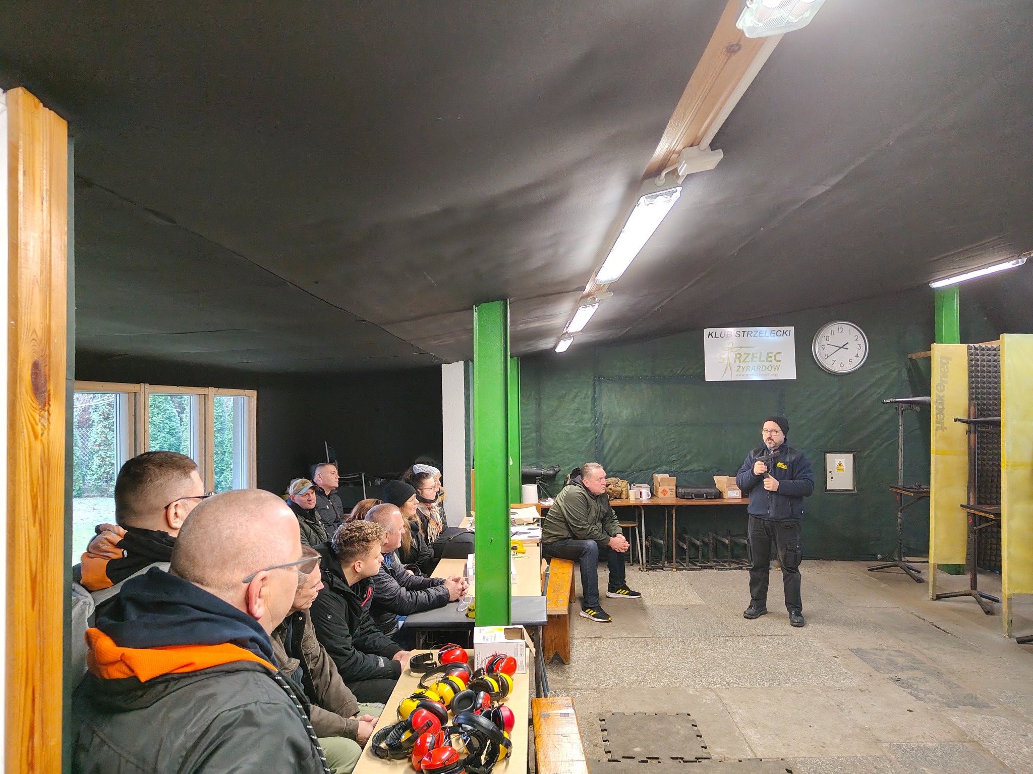 Instruktor prowadzi szkolenie strzeleckie na krytej strzelnicy. Uczestnicy siedzą przy stołach, na których leżą słuchawki ochronne. Widoczny zegar i logo klubu strzeleckiego.