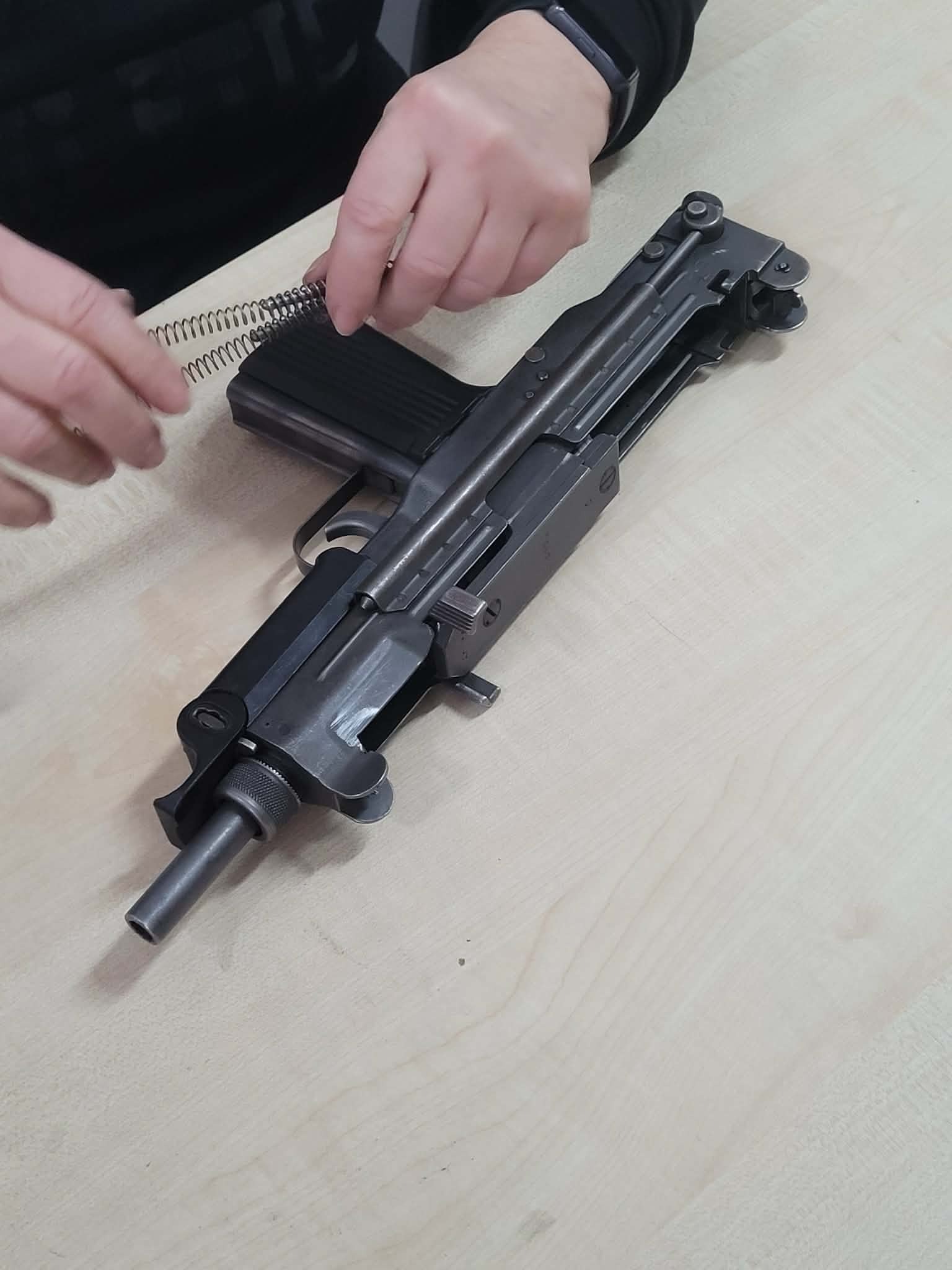 Rozłożony pistolet maszynowy UZI na jasnym stole. Dłonie wkładają sprężynę do magazynka. Widoczny mechanizm i elementy broni. Precyzyjne detale.