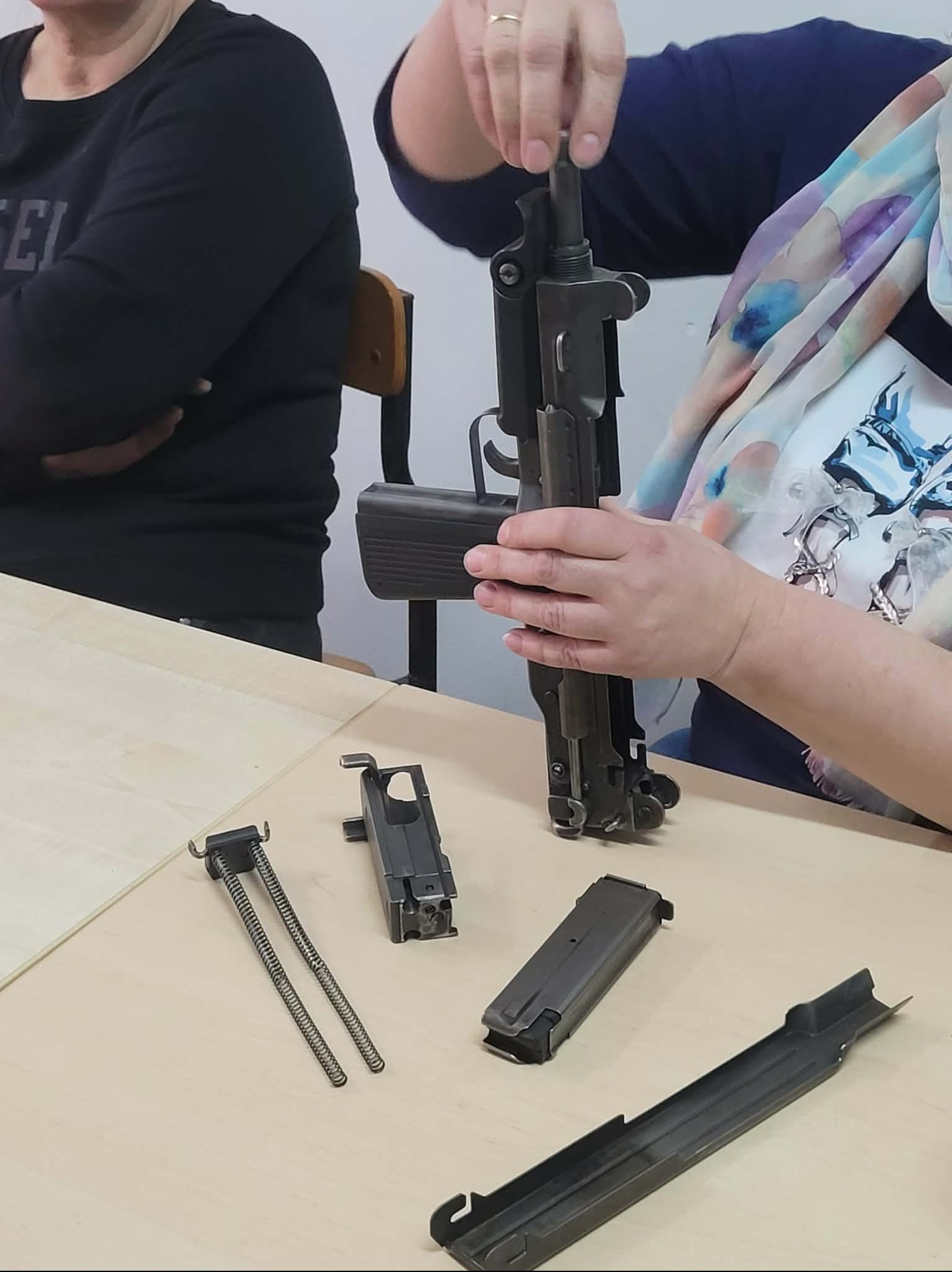 Rozłożony pistolet maszynowy UZI na stole, widoczne elementy mechanizmu, ręce osoby składającej broń. Szkolenie z obsługi broni palnej.
