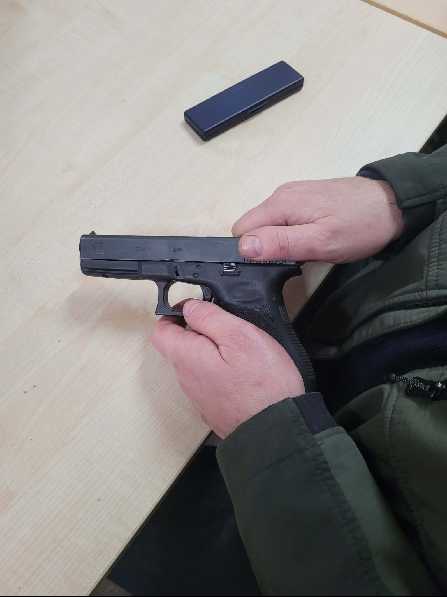 Czarny pistolet Glock 17 w dłoniach osoby na jasnym stole, obok czarny prostokątny przedmiot. Ujęcie z bliska, widoczna część zielonej kurtki.