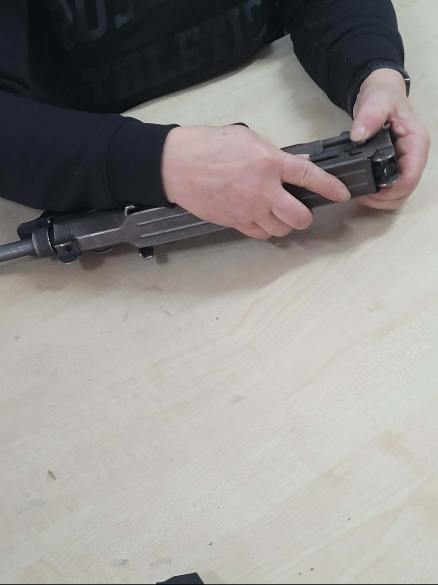 Instruktor prezentuje rozkładanie broni palnej na jasnym stole. Widoczne dłonie i fragment broni w trakcie demonstracji. Szkolenie z obsługi broni.