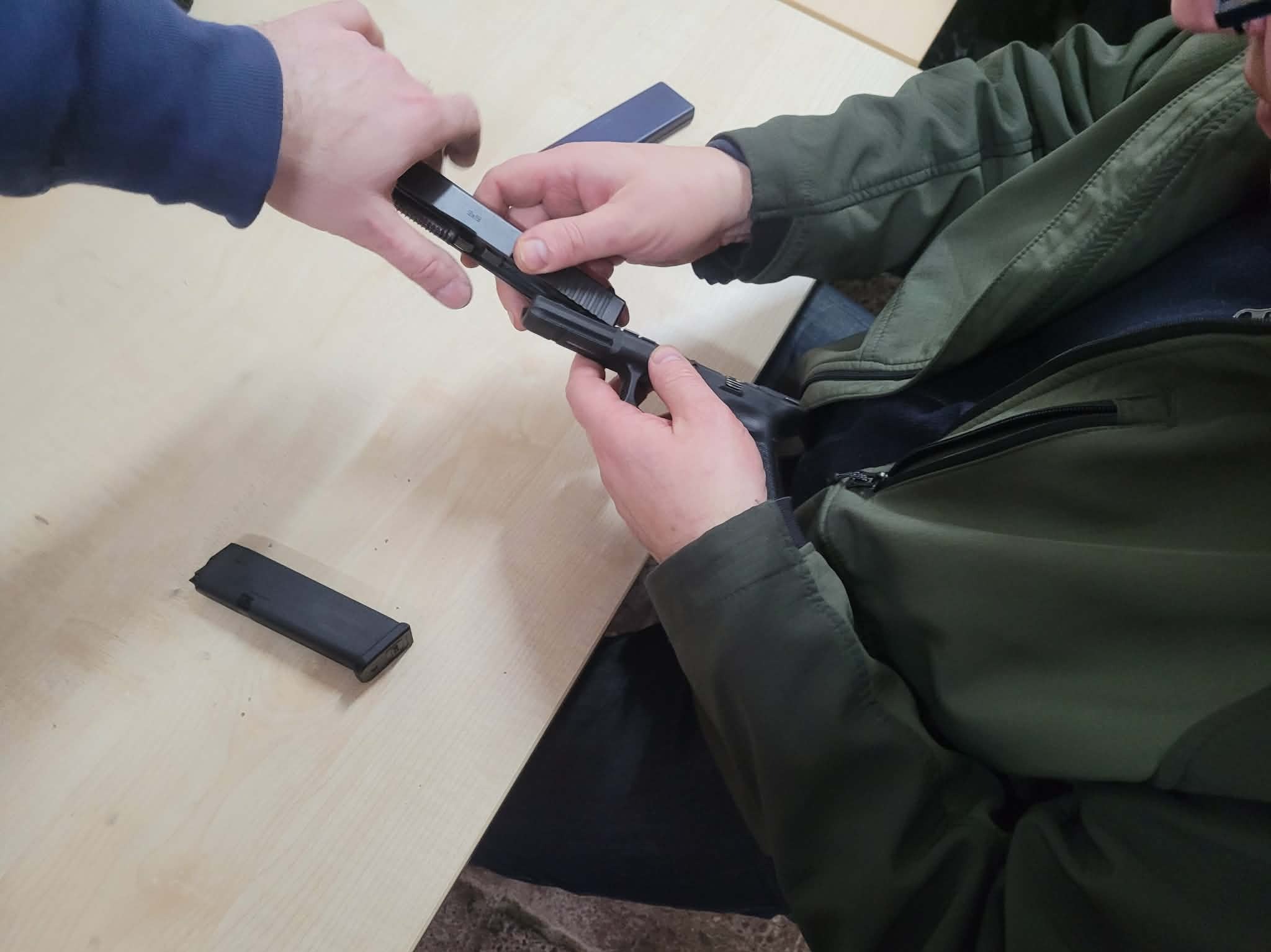Instruktorzy prezentują rozkładanie i składanie broni palnej Glock na jasnym stole podczas kursu strzeleckiego. Widoczny magazynek i dłonie uczestników.