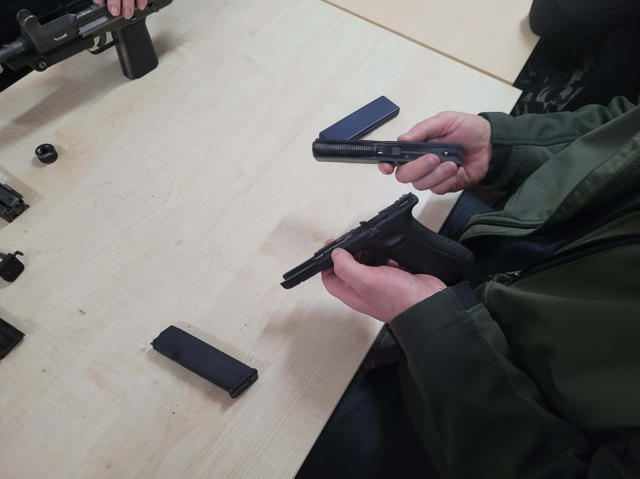 Rozłożony pistolet na stole, widoczne elementy broni i dłonie osoby, prawdopodobnie podczas szkolenia z obsługi broni palnej. Jasne oświetlenie, neutralne tło.