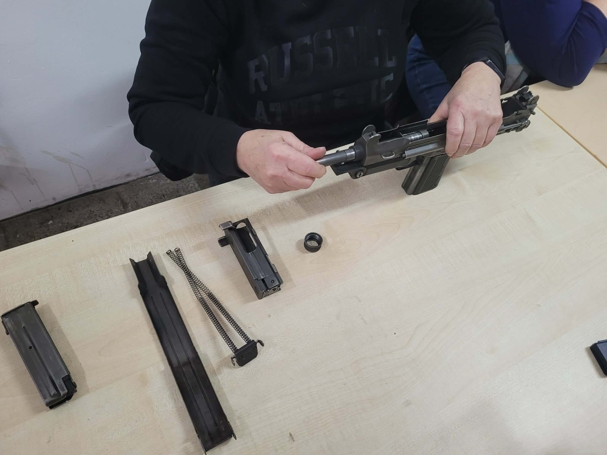 Demontaż broni palnej na stole: osoba wkłada element do karabinu, obok leżą magazynki i sprężyny. Szkolenie z obsługi broni.
