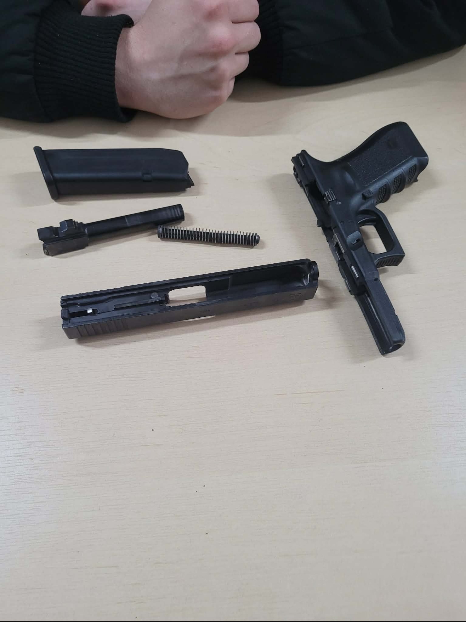 Rozłożony pistolet Glock na jasnym stole, widoczne elementy: magazynek, zamek, lufa i sprężyna. Dłoń w tle. Prezentacja budowy i mechanizmów broni palnej.