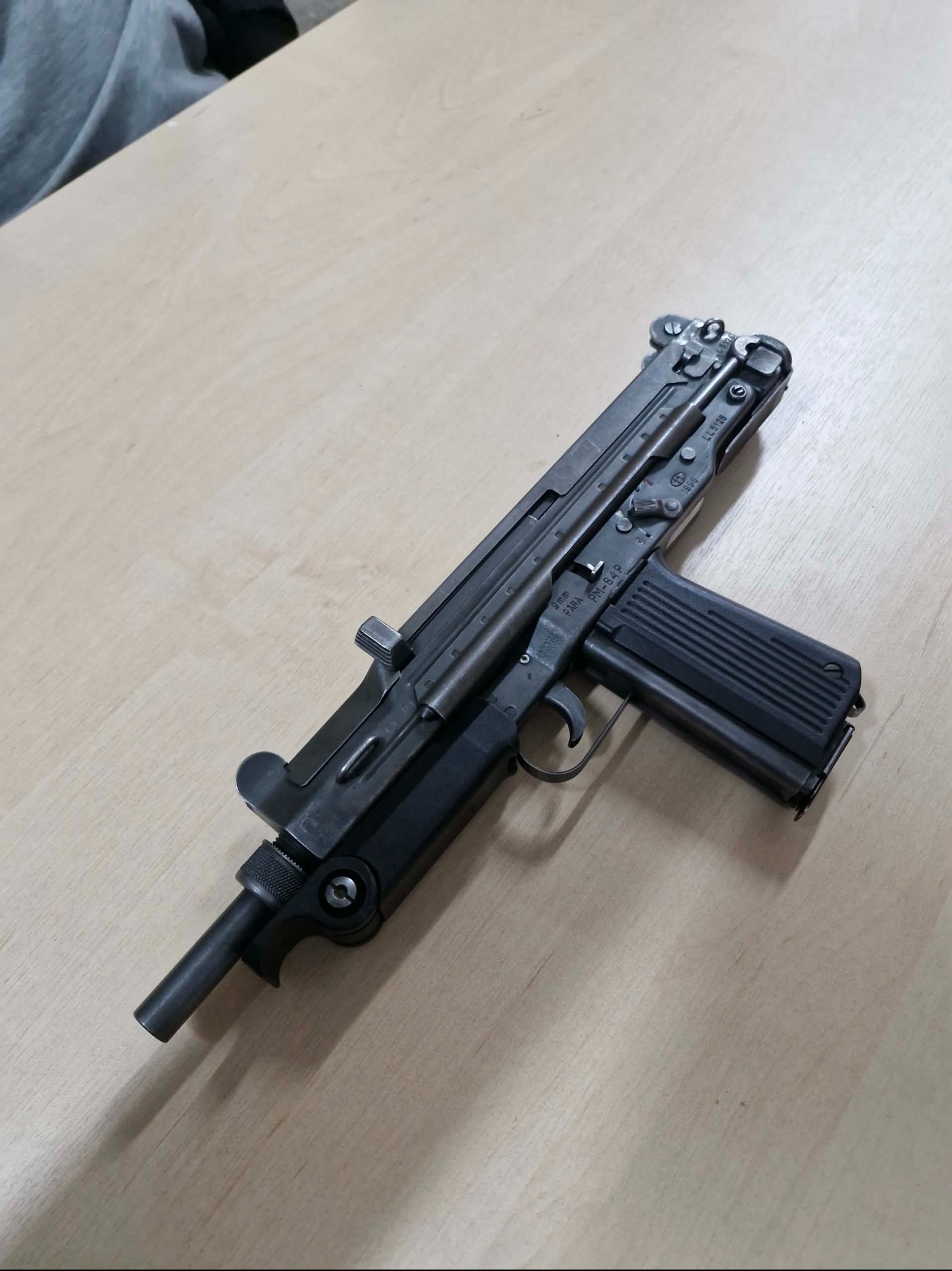 Pistolet maszynowy PM-84P FARA kaliber 9mm, leżący na jasnym drewnianym stole. Widoczny magazynek i elementy mechaniczne. Ujęcie z góry, oświetlenie naturalne.