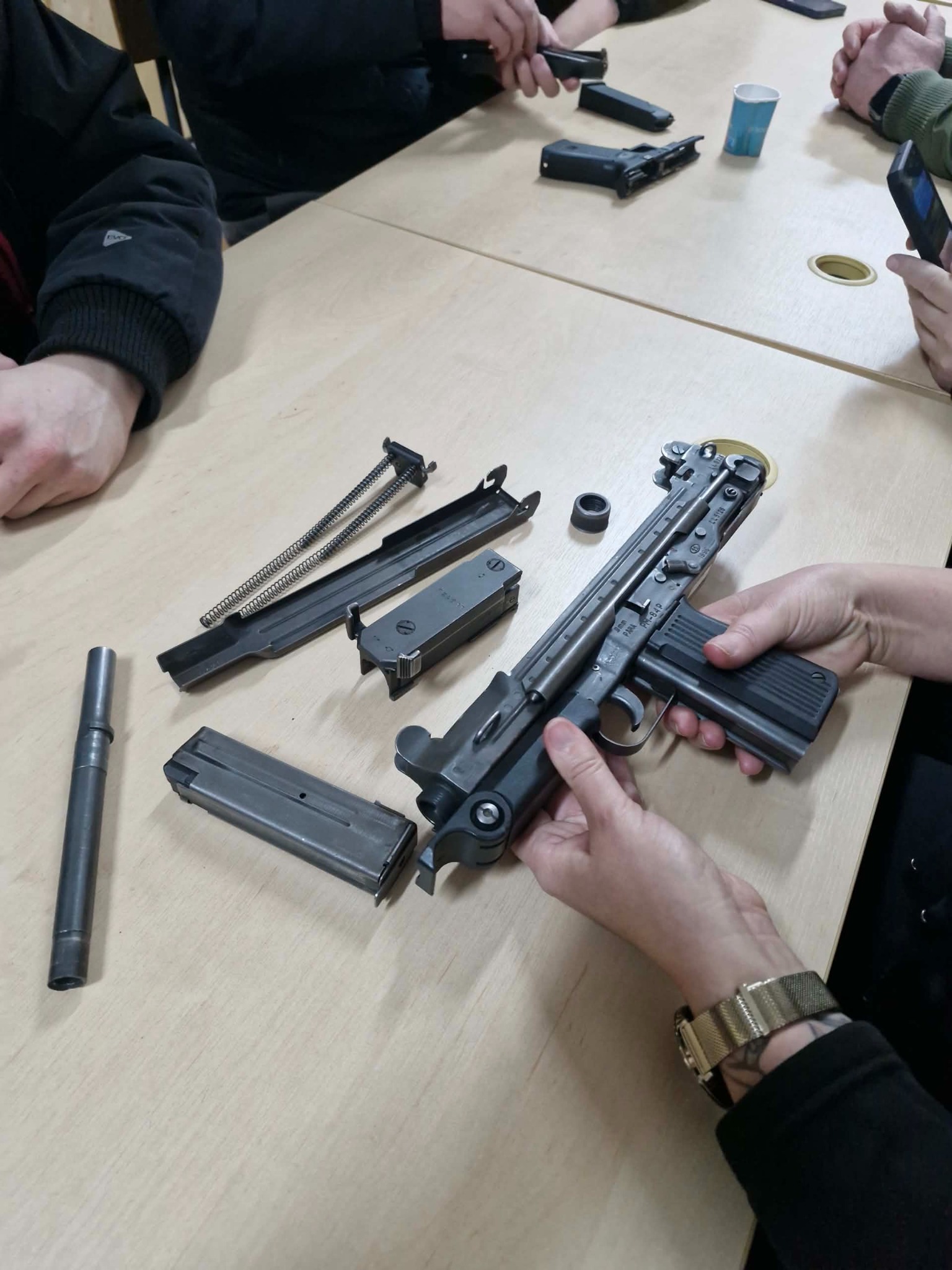 Rozłożony pistolet maszynowy PPS wz. 43 na stole podczas szkolenia. Widoczne elementy broni i dłonie uczestników kursu. Detaliczny widok na mechanizm i magazynek.