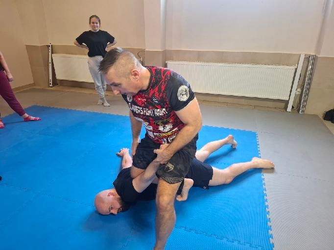 Instruktor prezentuje technikę walki na macie podczas kursu samoobrony. Uczestniczka obserwuje z tyłu. Oświetlenie dzienne, sala treningowa.