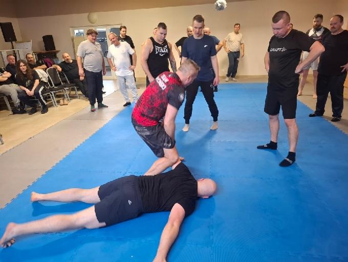 Sala treningowa: mężczyzna w czarnej koszulce leży na niebieskiej macie, drugi demonstruje technikę, grupa obserwuje. Trening samoobrony.