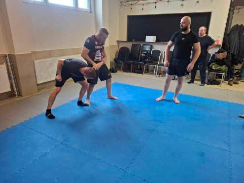 Trening sztuk walki na niebieskiej macie: dwóch mężczyzn w zwarciu, trzeci obserwuje. Sala z krzesłami i tablicą w tle. Ujęcie z boku, naturalne światło.