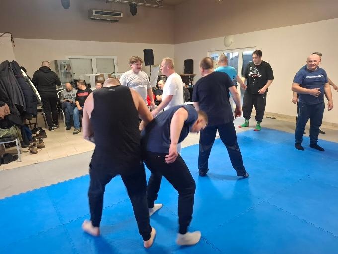 Sala treningowa z grupą osób ćwiczących techniki samoobrony na niebieskiej macie. Widoczni instruktorzy i uczestnicy w trakcie zajęć.