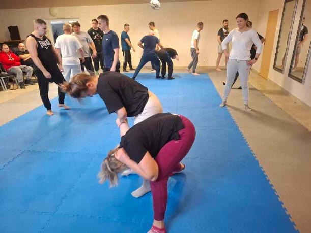 Sala treningowa z grupą osób ćwiczących techniki samoobrony na niebieskiej macie. Instruktor nadzoruje ćwiczenia. Widoczne lustra na ścianie.