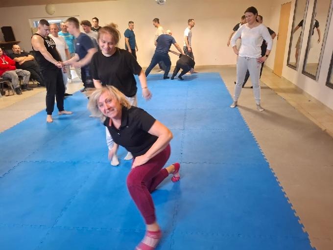 Sala treningowa z grupą osób ćwiczących techniki samoobrony na niebieskiej macie. Instruktorzy demonstrują chwyty, a uczestnicy powtarzają ruchy.