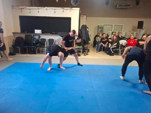 Trening sztuk walki na niebieskiej macie. Dwóch mężczyzn w czarnych strojach w trakcie sparingu, publiczność obserwuje z krzeseł w tle.