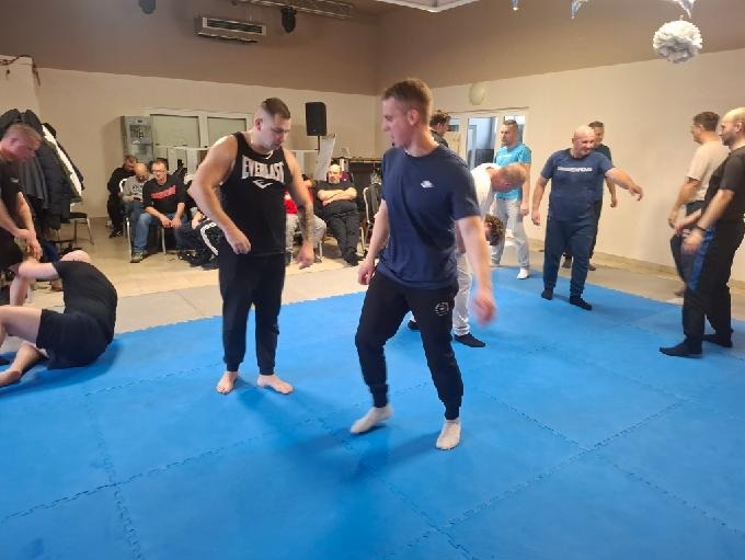 Sala treningowa z grupą mężczyzn ćwiczących sztuki walki na niebieskiej macie. Widoczni instruktorzy i uczestnicy w trakcie zajęć, obserwujący siedzą na krzesłach w tle.