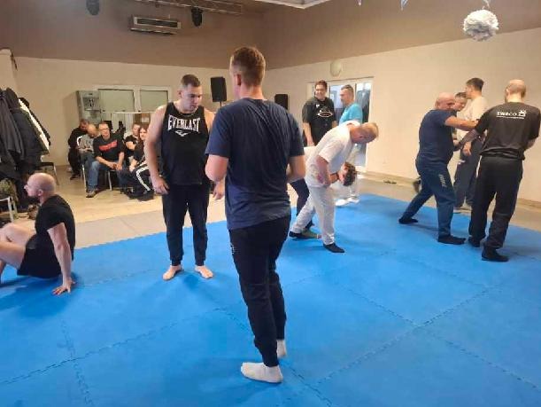 Grupa mężczyzn ćwiczy na niebieskiej macie w sali. Instruktorzy demonstrują techniki, a kursanci powtarzają. Widoczni obserwatorzy i elementy wyposażenia sali.