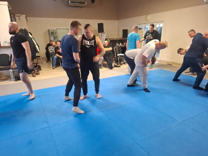 Trening samoobrony: grupa mężczyzn ćwiczy techniki walki na niebieskiej macie w sali. Widoczni obserwatorzy i wyposażenie sali treningowej.