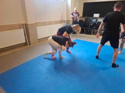 Trening samoobrony w Skierniewicach: grupa osób ćwiczy techniki walki na niebieskiej macie w sali. Instruktor nadzoruje ćwiczenia.