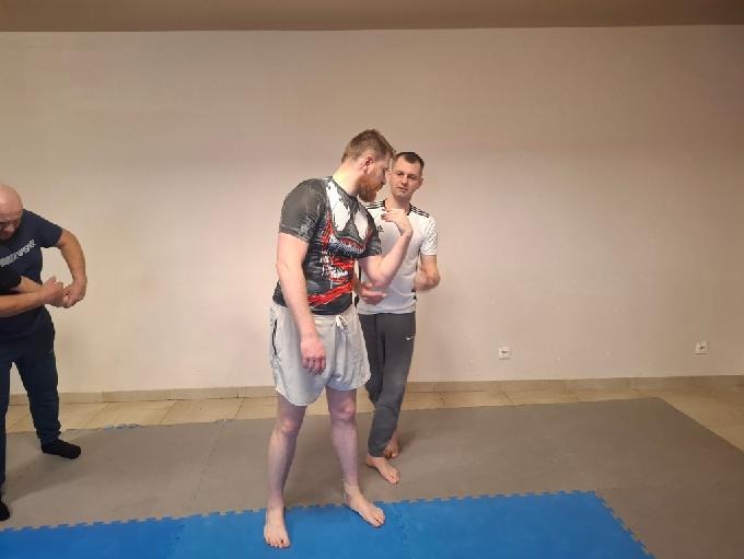 Instruktor demonstruje technikę samoobrony na kursancie podczas zajęć na macie. Widoczni inni uczestnicy szkolenia.