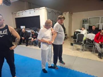 Instruktor demonstruje technikę samoobrony na uczestniku kursu w sali szkoleniowej. Widoczni inni kursanci i wyposażenie sali.