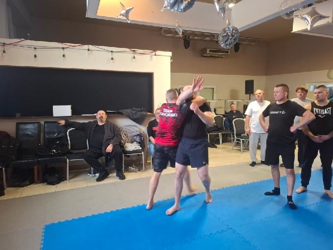 Trening samoobrony: dwóch mężczyzn w zwarciu na niebieskiej macie, grupa obserwatorów w tle. Sala z dekoracjami.