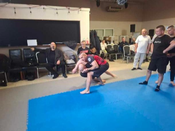 Trening samoobrony: dwóch mężczyzn w trakcie ćwiczenia chwytów na niebieskiej macie, obserwatorzy w tle. Sala z czarną tablicą i krzesłami.