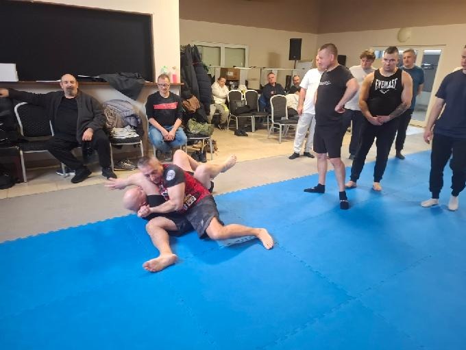 Trening sztuk walki na niebieskiej macie: dwóch mężczyzn w zwarciu, grupa obserwatorów w tle. Sala z krzesłami i czarną tablicą. Oświetlenie sufitowe.