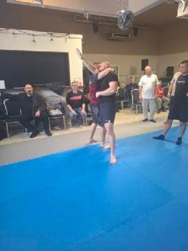 Trening samoobrony: dwóch mężczyzn w zwarciu na niebieskiej macie, obserwujących kilku mężczyzn w tle. Sala z oświetleniem sufitowym i krzesłami.