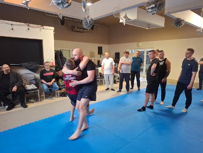 Trener w czarnej koszulce obejmuje ucznia podczas sparingu na niebieskiej macie. W tle obserwujący kursanci i dekoracje sali.