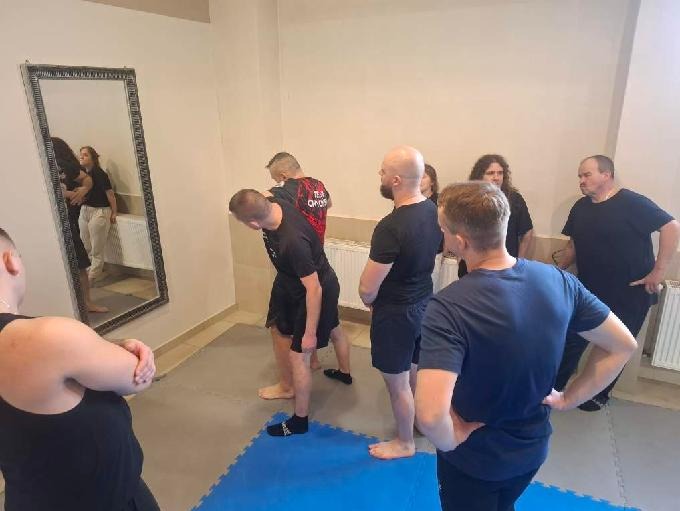 Grupa osób w czarnych strojach ćwiczy w sali z lustrem i niebieską matą. Jeden z uczestników ma koszulkę z napisem 'TEAM ORION'.