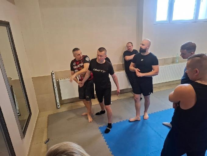 Sala treningowa: instruktor demonstruje chwyt na ręce uczestnikowi kursu samoobrony. Grupa obserwuje, w tle lustro i grzejnik.