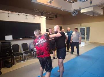 Instruktor demonstruje technikę samoobrony na sali treningowej. Grupa obserwatorów w tle. Widoczna tablica i sprzęt treningowy. Oświetlenie sufitowe.