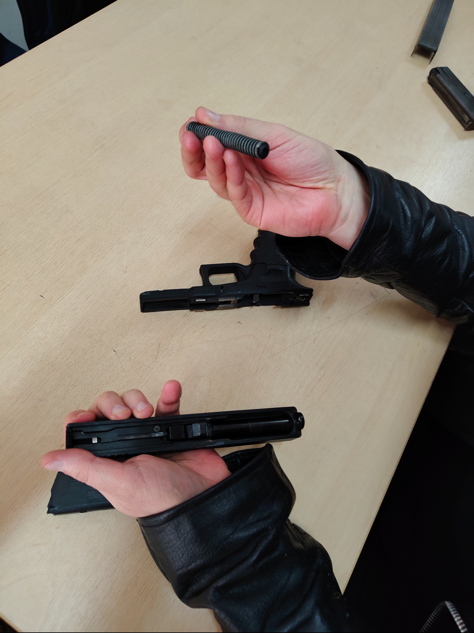 Rozłożony pistolet Glock na stole, widoczne elementy: szkielet, zamek, sprężyna powrotna. Osoba w czarnej kurtce trzyma sprężynę. Precyzyjne ujęcie detali mechanizmu.