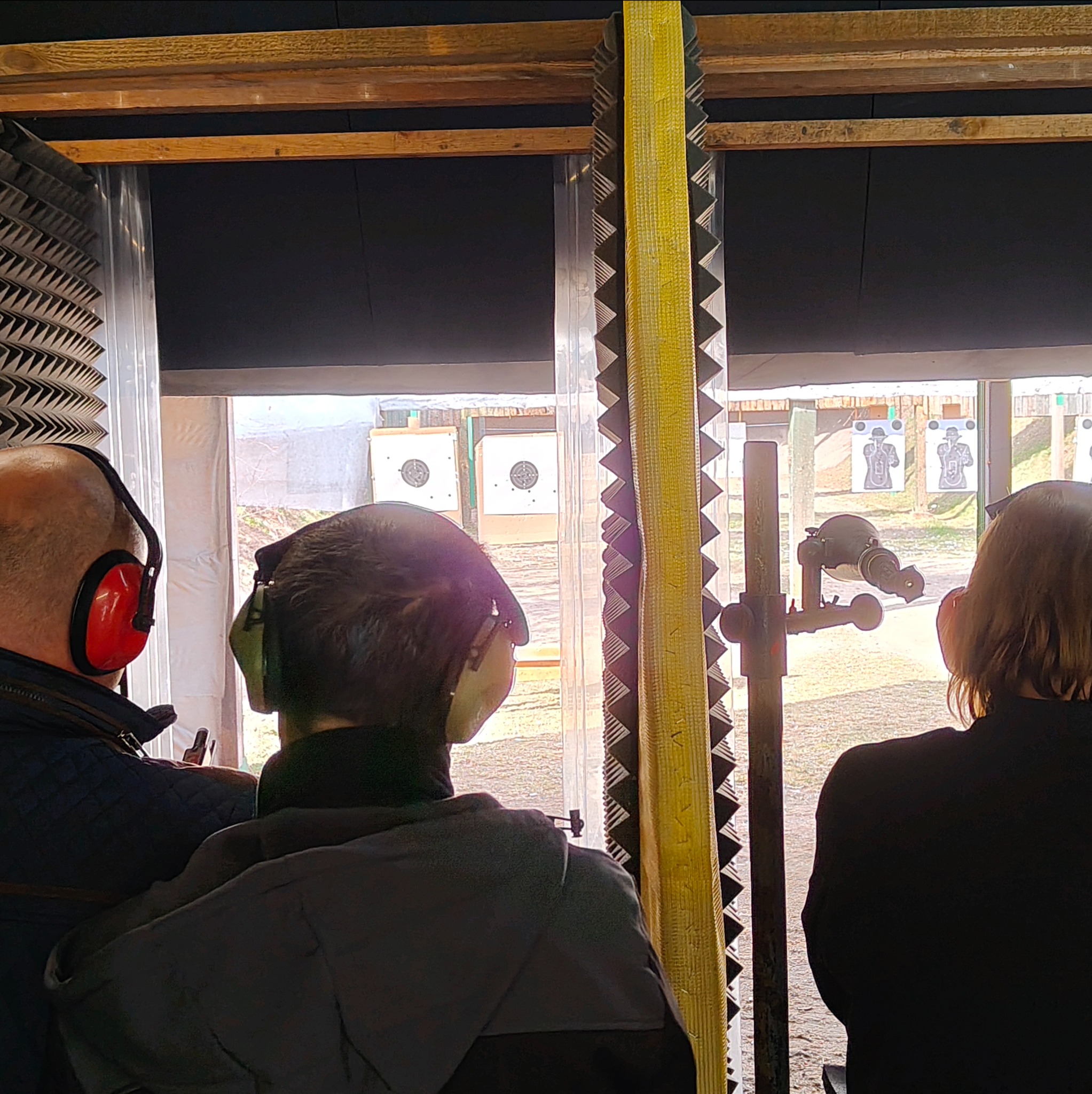 Osoby w słuchawkach ochronnych na strzelnicy, celują do tarcz. Widoczne tarcze z sylwetkami i okrągłe. Elementy wygłuszające na ścianach. Nauka strzelania.