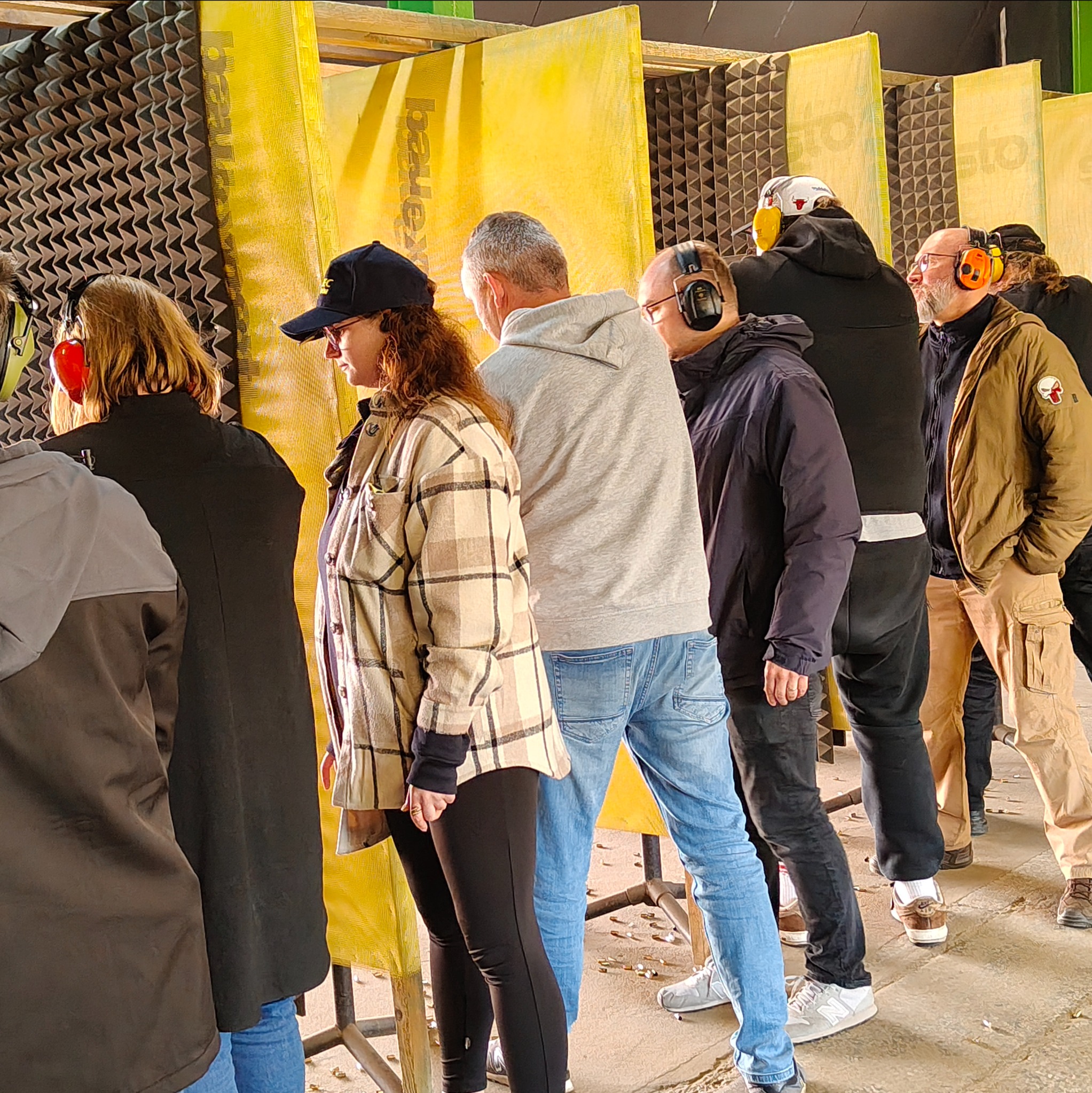 Grupa osób w słuchawkach ochronnych na strzelnicy, widoczne żółte przegrody i łuski nabojów na podłodze. Uczestnicy kursu strzeleckiego podczas zajęć praktycznych.