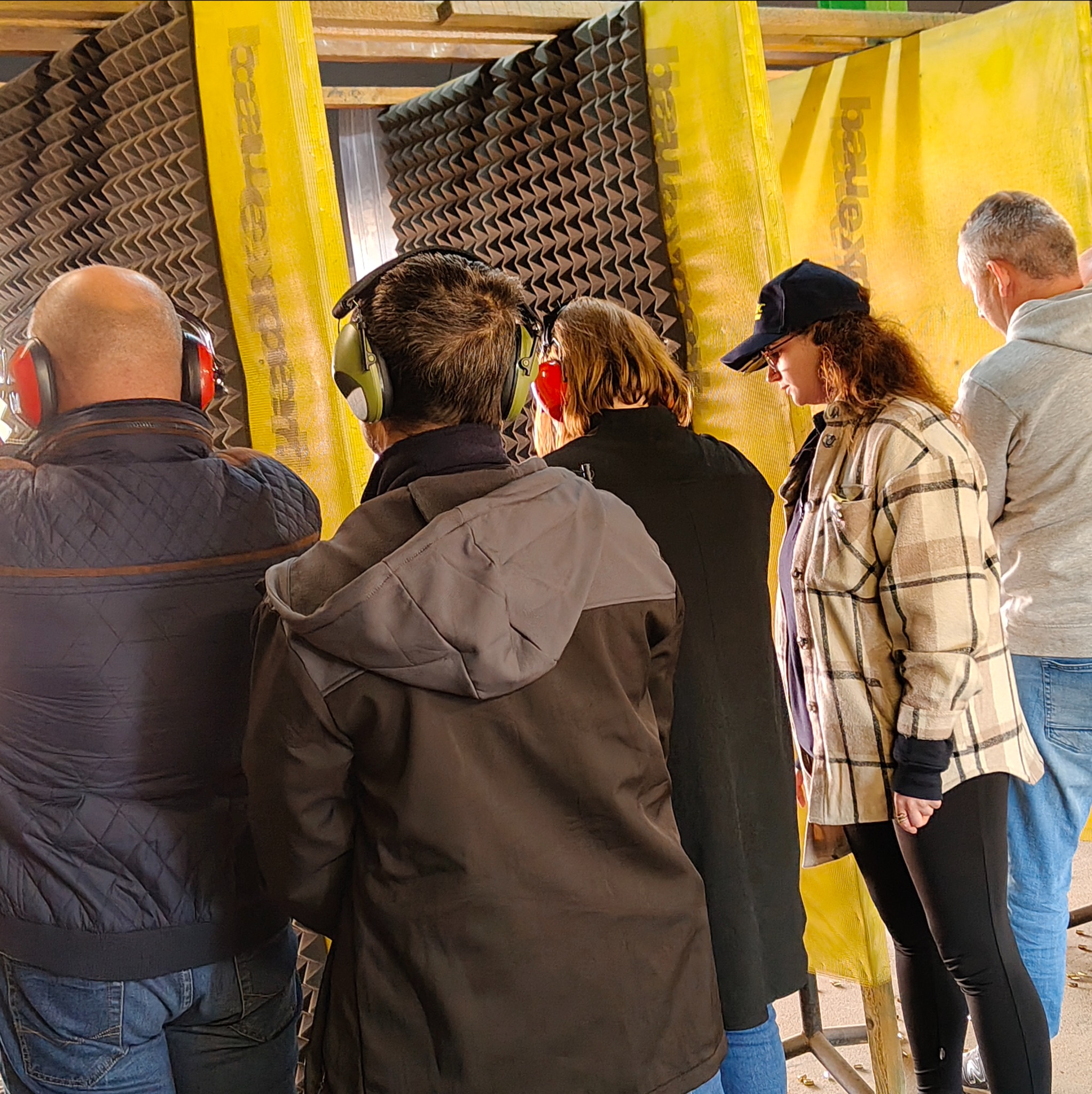 Grupa osób w słuchawkach ochronnych na strzelnicy, z widocznymi żółtymi panelami dźwiękochłonnymi i łuskami nabojów na podłodze. Uczestnicy kursu strzeleckiego.