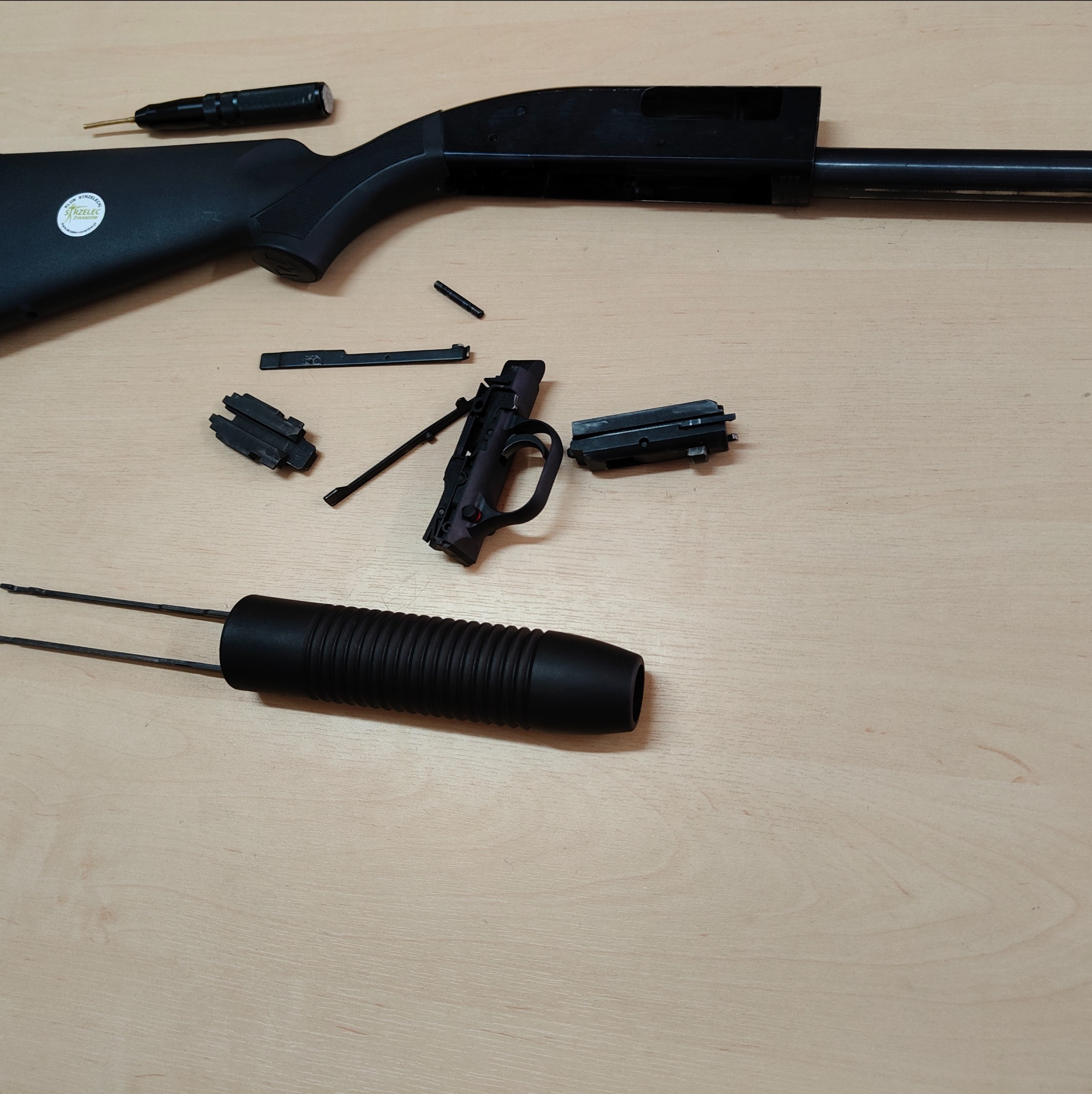 Rozłożona czarna strzelba na drewnianym stole. Widoczne elementy: kolba z naklejką, lufa, uchwyt, sprężyna i inne części. Prezentacja demontażu broni.