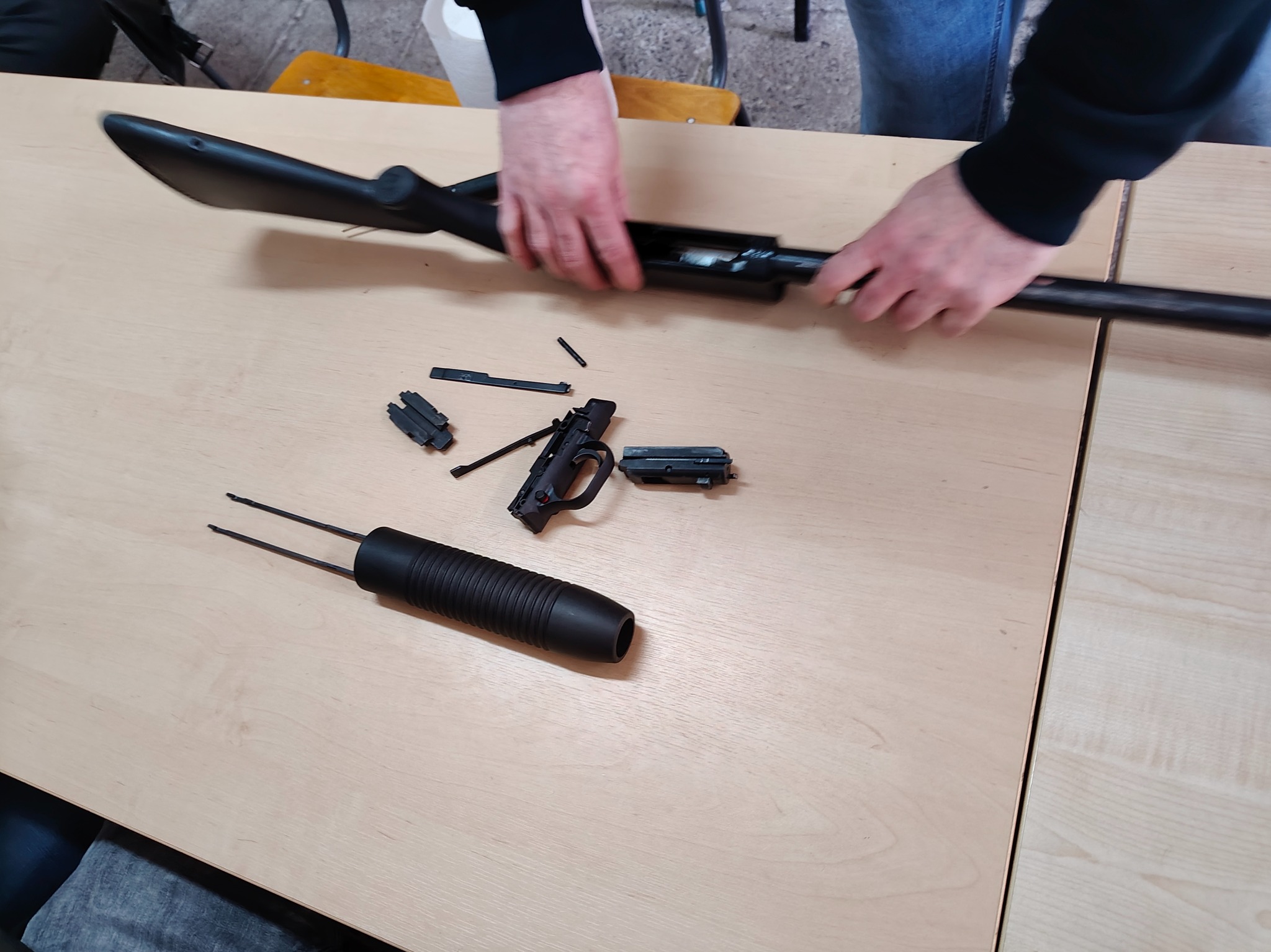 Rozłożona broń palna na stole podczas szkolenia z obsługi. Widoczne elementy broni i dłonie osoby składającej. Jasne oświetlenie, neutralne tło.