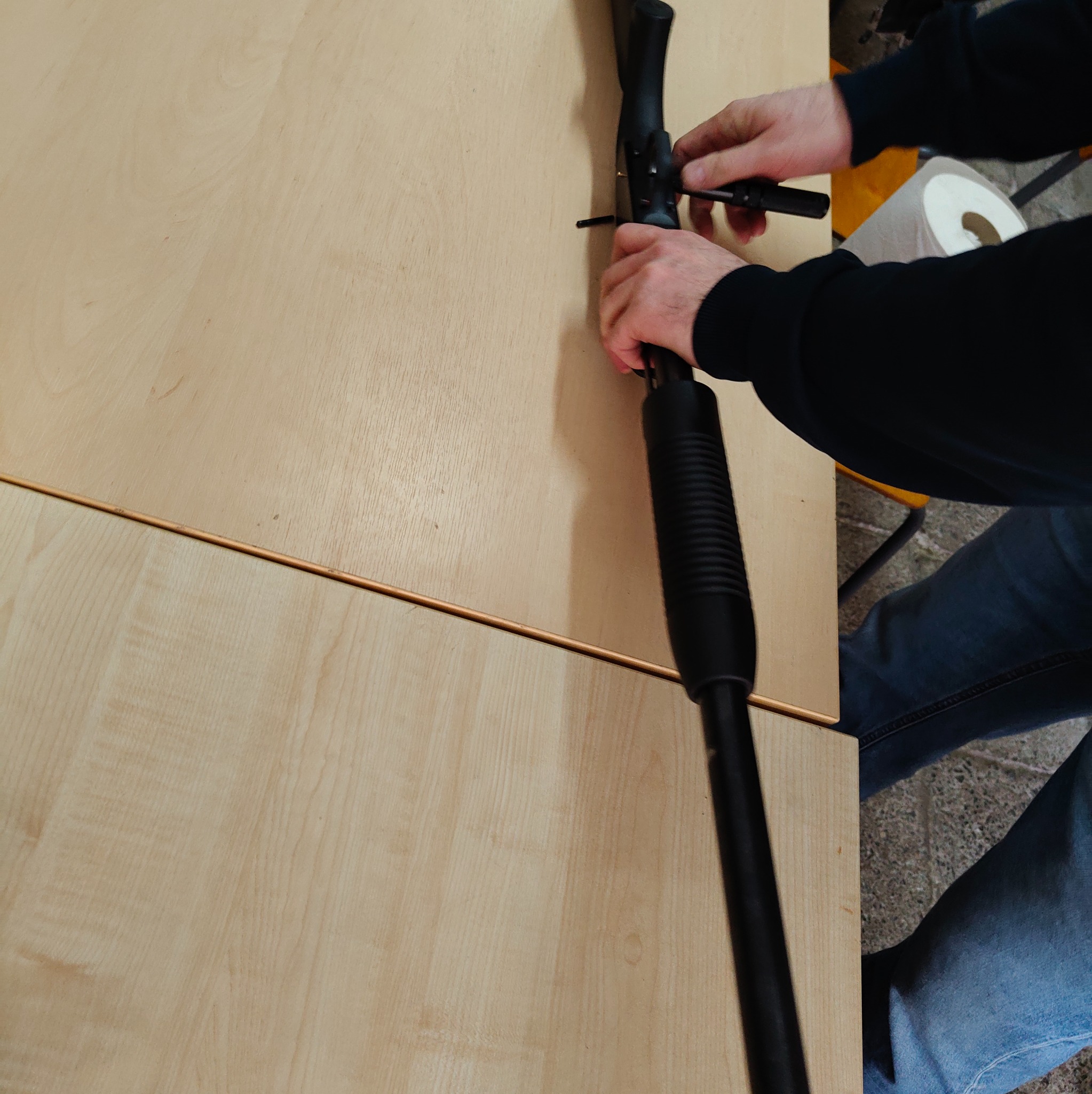 Czyszczenie broni palnej na drewnianym stole. Widoczne dłonie, fragment broni, narzędzie do czyszczenia i rolka papieru. Ujęcie z góry, oświetlenie naturalne.